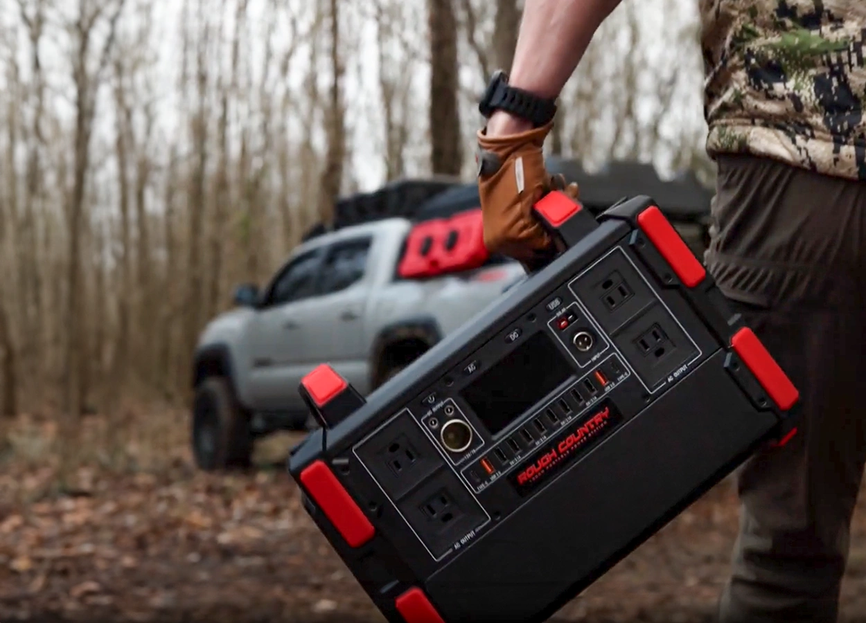 A man in a wooded area wearing camo carrying a Rough Country Portable Power Station as he walks towards his off-road truck