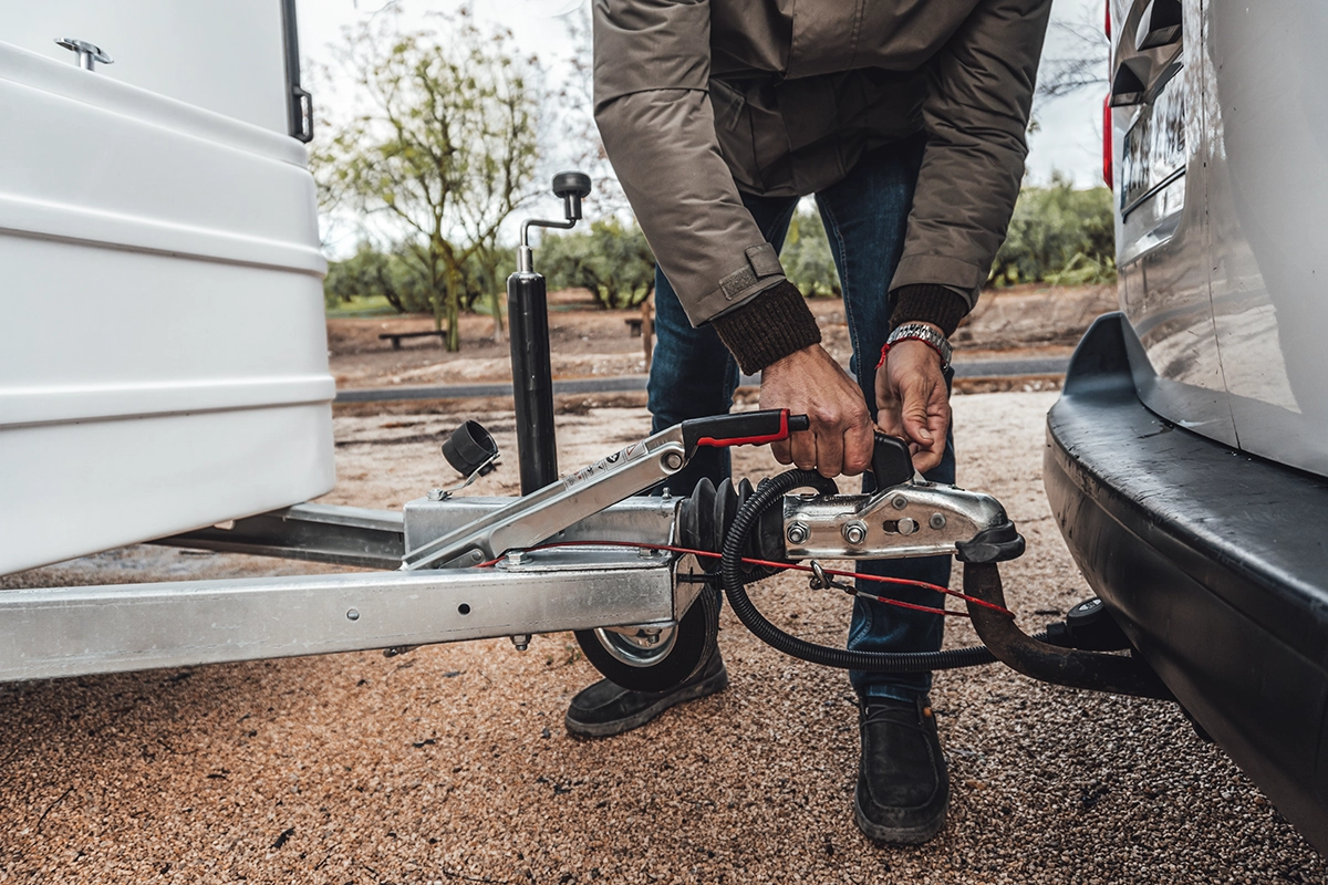 A pickup truck driver adjusts the trailer hitch attached to a white truck and a trailer on the side of the road with trees in the background, showing the concept of towing accessories