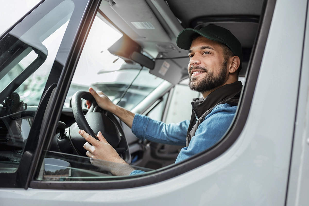 A smiling commercial fleet driver at the wheel of a work truck or van, wearing a hat and blue jacket