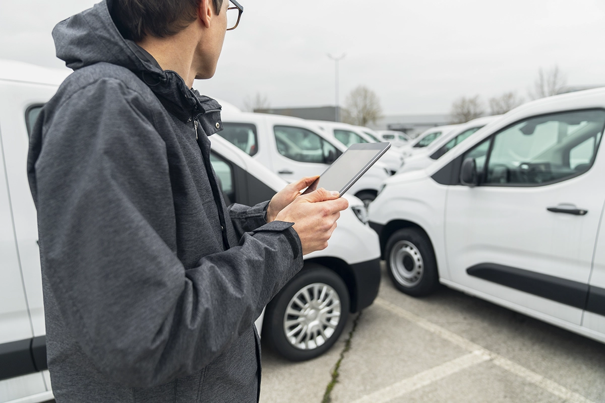 A commercial fleet manager in a dark gray jacket holds a tablet, inspecting a parking lot of work vans, showing the concept of vehicle inspections