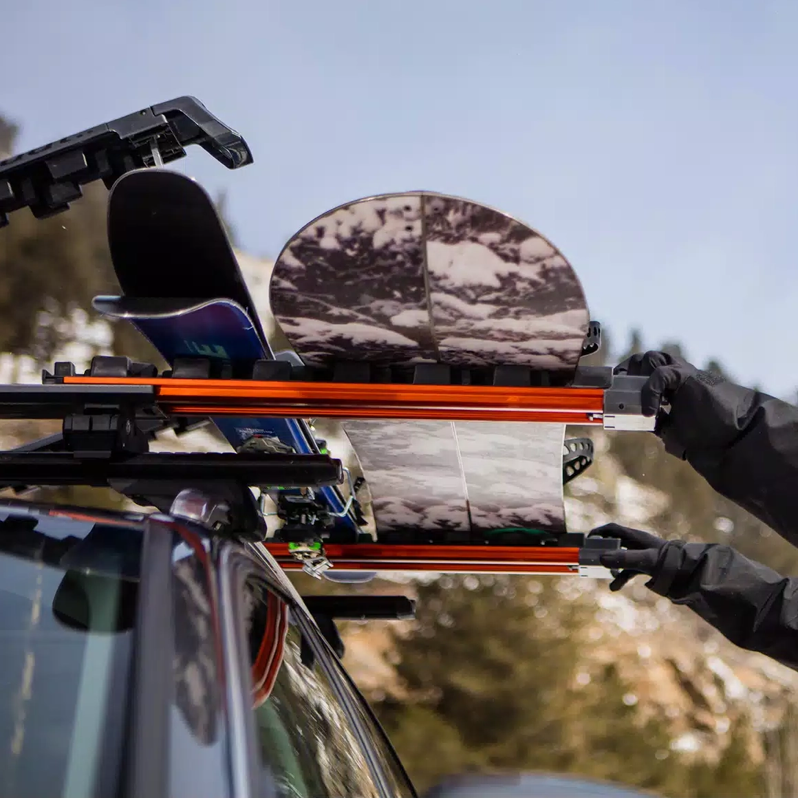 A Kuat Grip ski and snowboard rack mounted to the roof of an SUV, with mountains in the background
