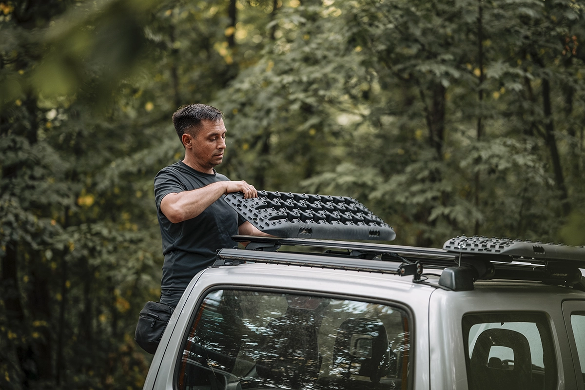 An overlander grabbing black traction boards from a silver off-roading vehicle, with green trees and a forest in the background, showing the concept of recovery gear for overlanding.