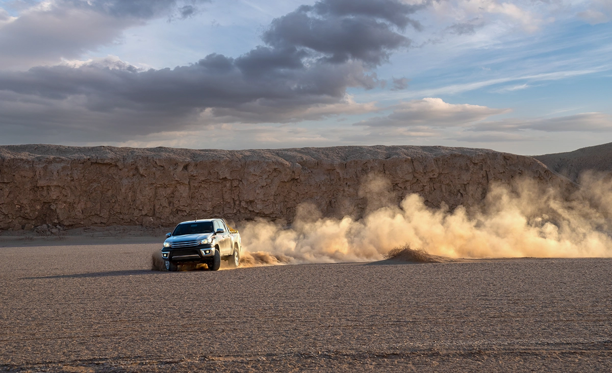 A white pickup truck off-roading in the desert at sunset, leaving a sand cloud trail behind it