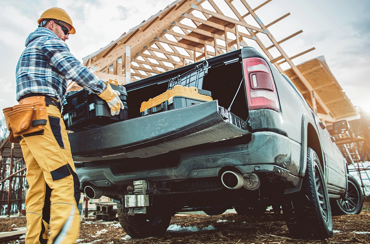 A construction worker in flannel and a yellow hard hat loads a payload of heavy tools into the bed of a dark gray pickup truck