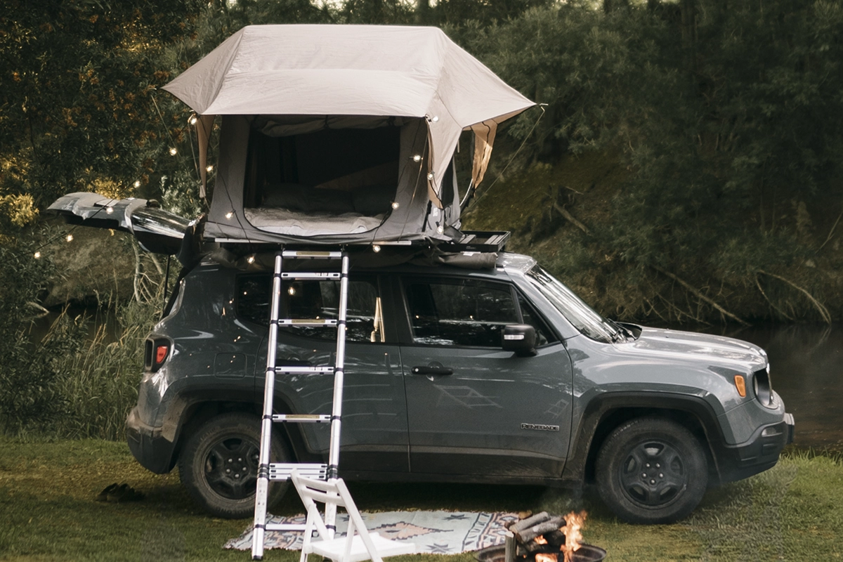 A rooftop tent on a blue-gray SUV, in a forest, next to a pond