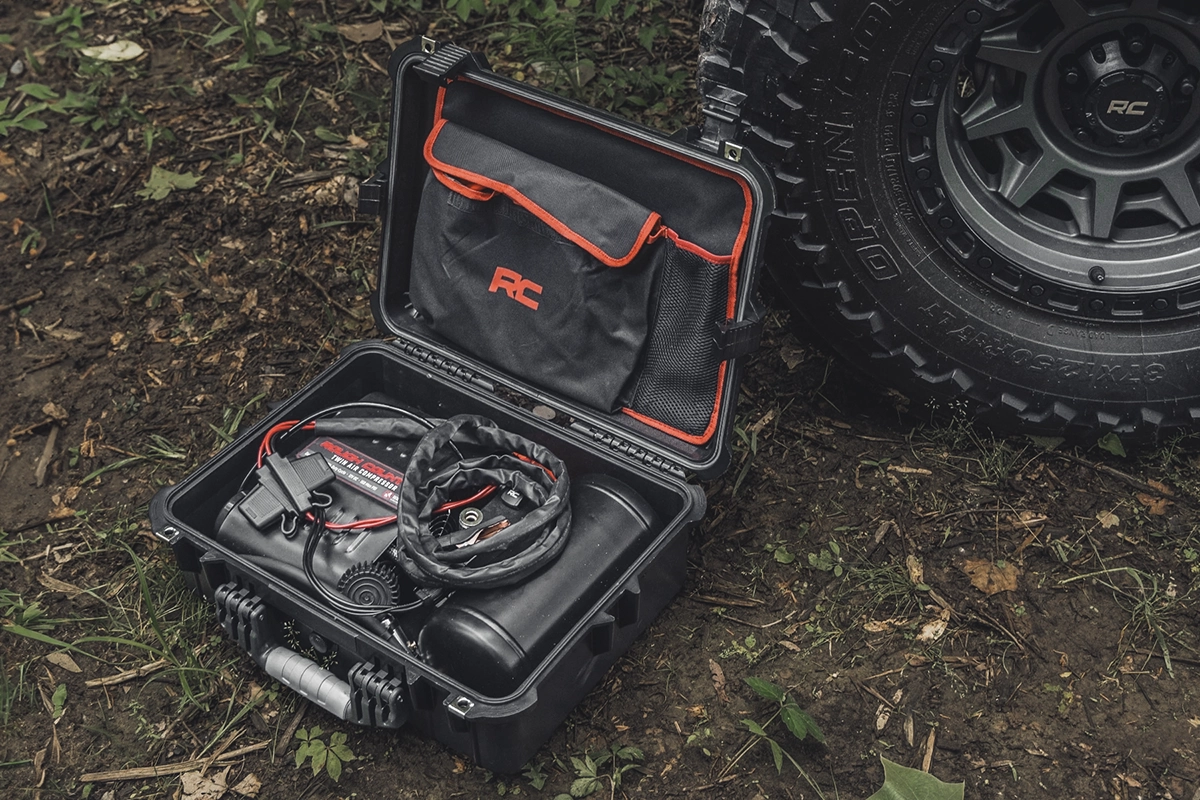 A Rough Country twin motor tire air compressor kit in a black carrying case, next to a tire, showing the concept of recovery gear for off-roading and overlanding