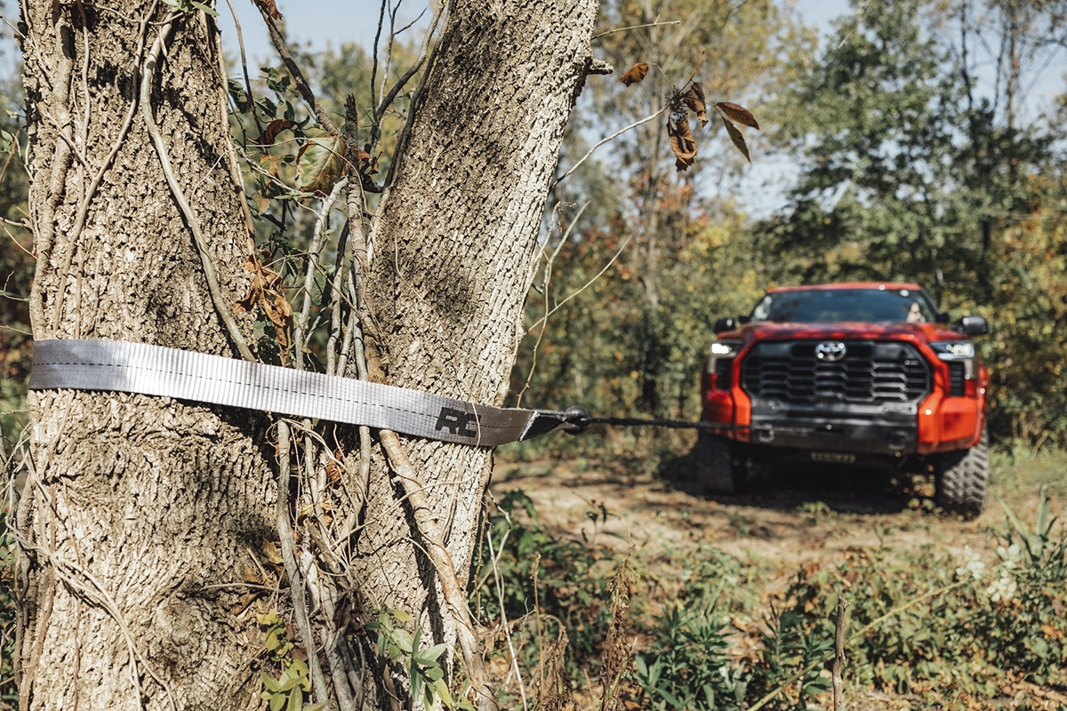 A red off-roading pickup truck using a tree saver strap and a winch to get unstuck in the woods, showing the concept of overlanding recovery gear