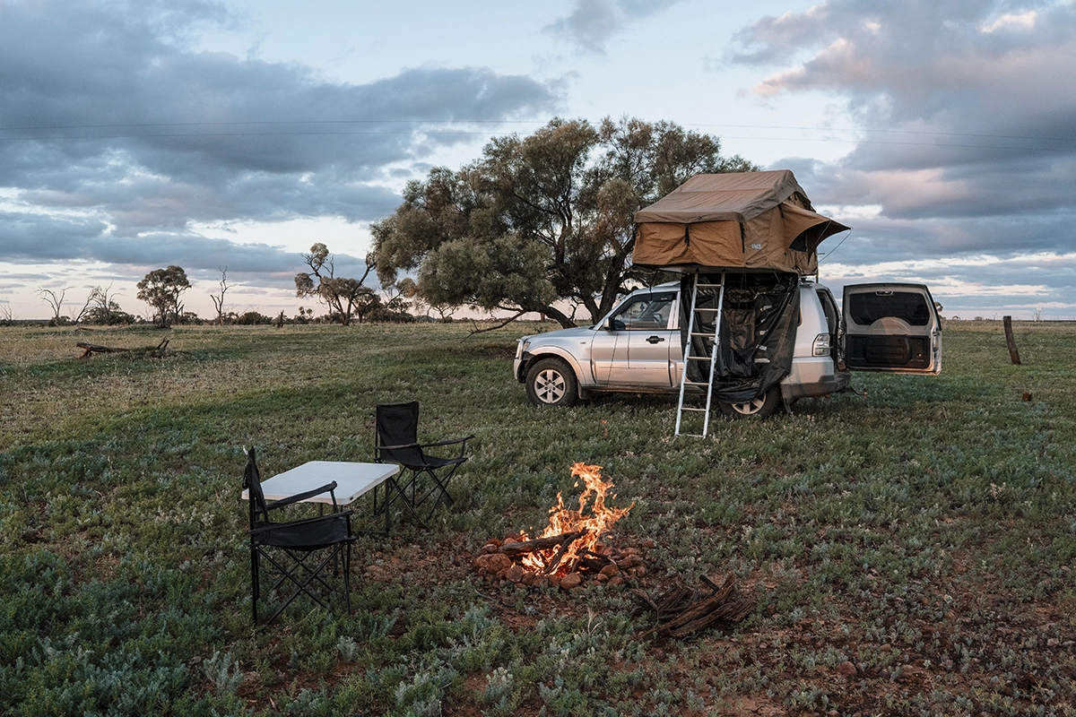 A silver SUV in a field of grass with a rooftop tent, at a campsite with a fire and folding chairs