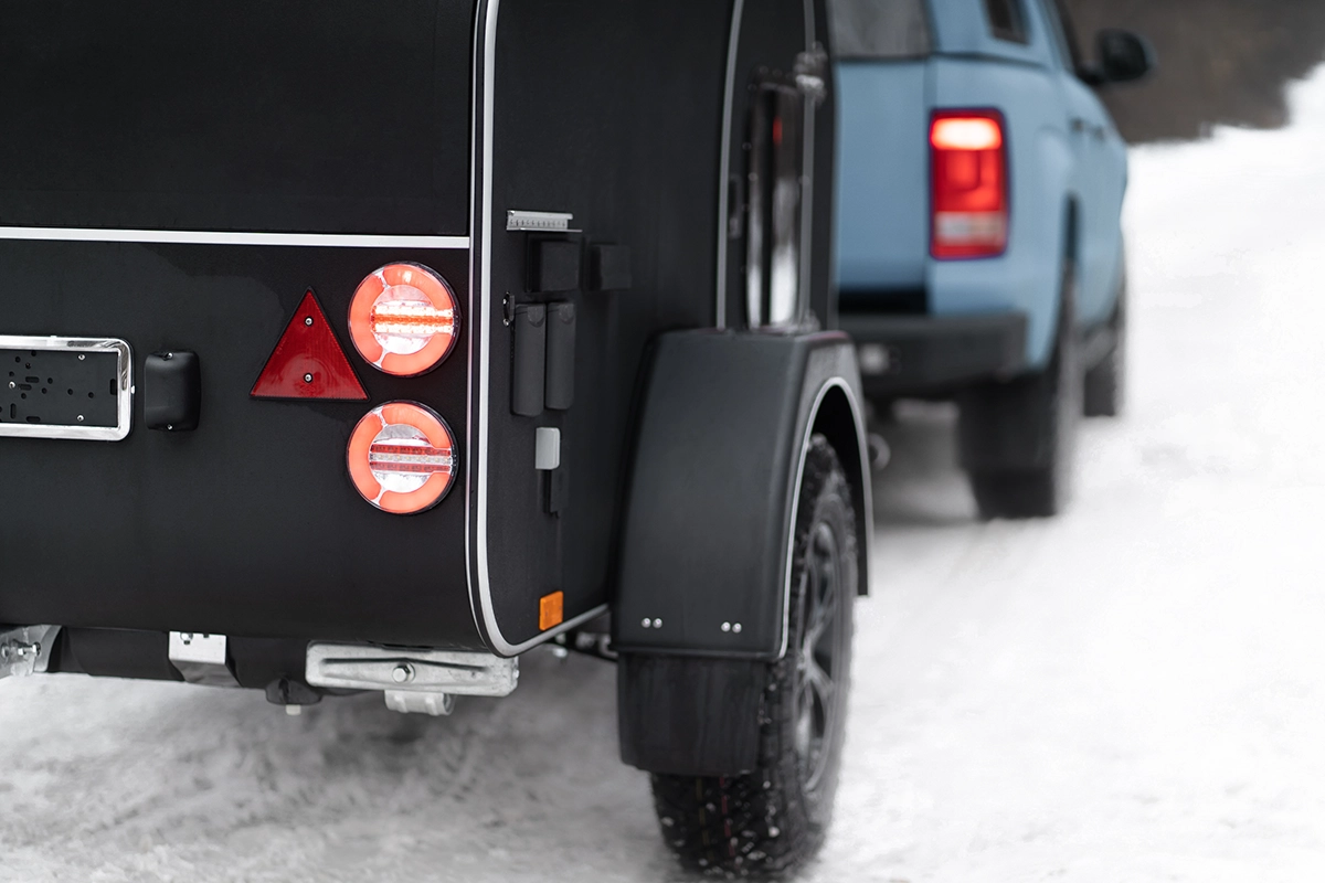 A close-up of trailer braking lights turned on, on the back of a black trailer, being towed by a blue pickup truck in the snow