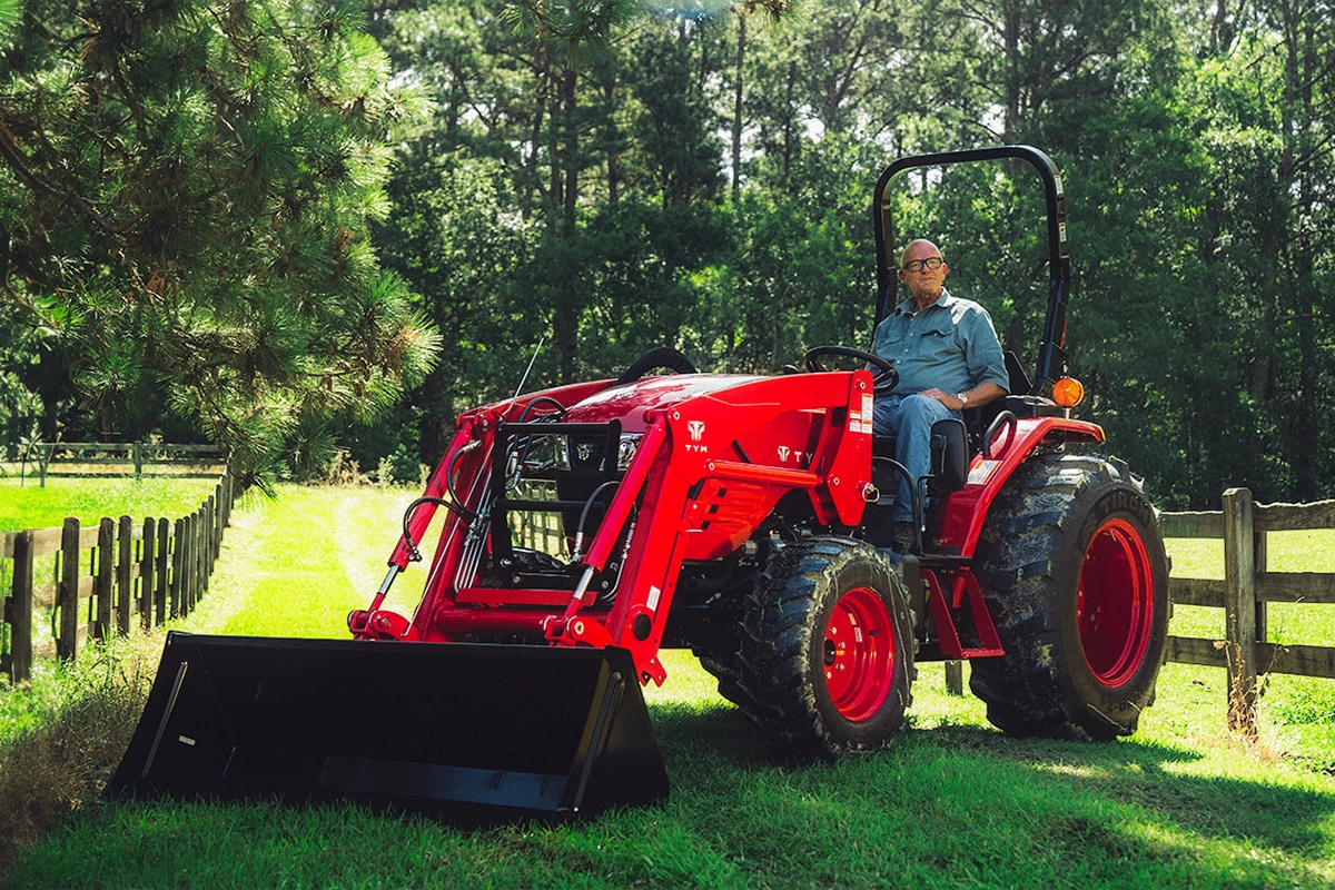 A man sitting on a red TYM Series 3 compact tractor in a field, surrounded by trees
