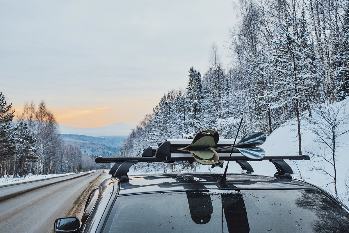 An SUV driving on a snowy mountain surrounded by trees at sunset, with a ski or snowboard winter sports rack mounted to the roof.