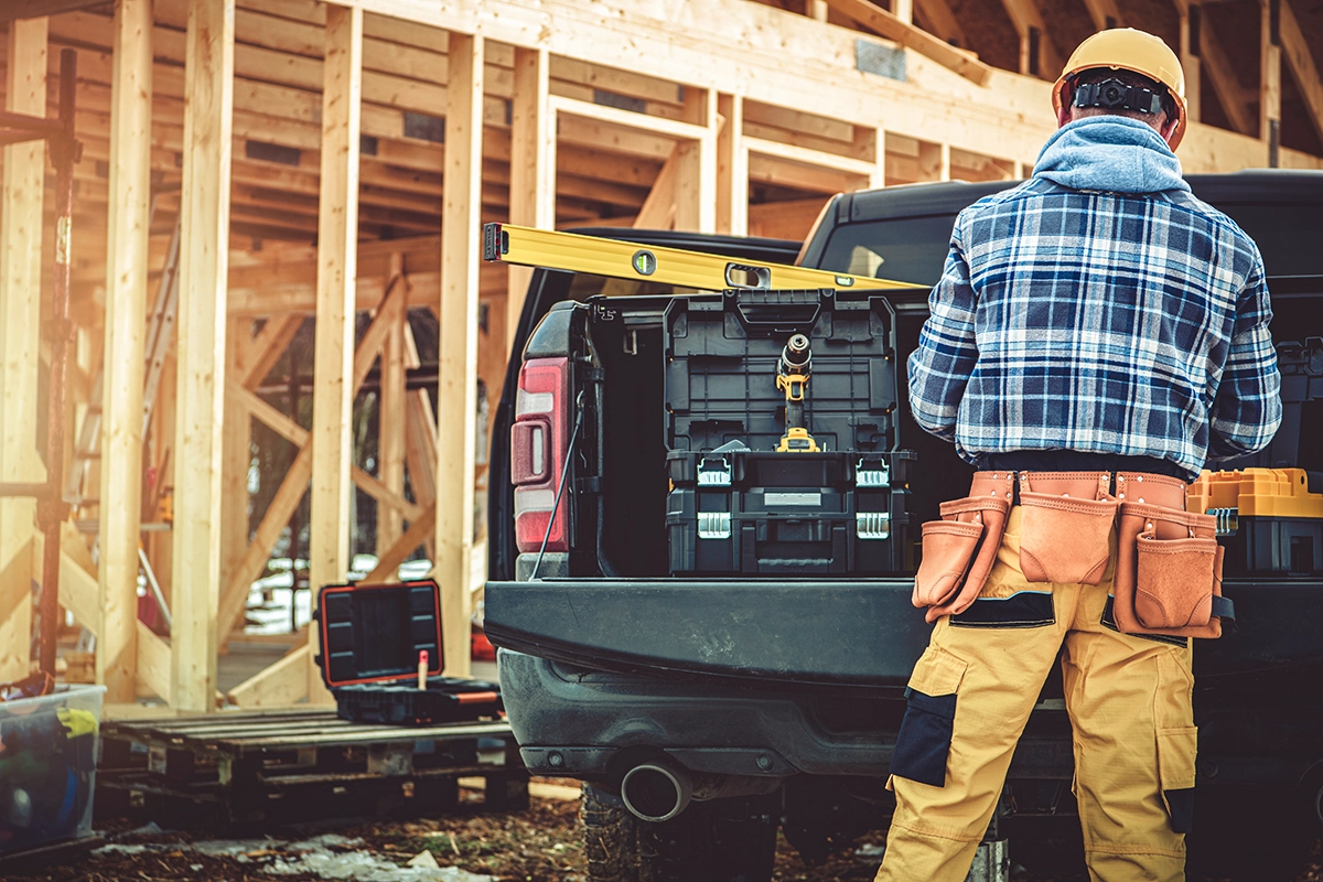 The back of a construction worker wearing a blue flannel and a yellow hard hat, getting something from the back of their work truck at a job site