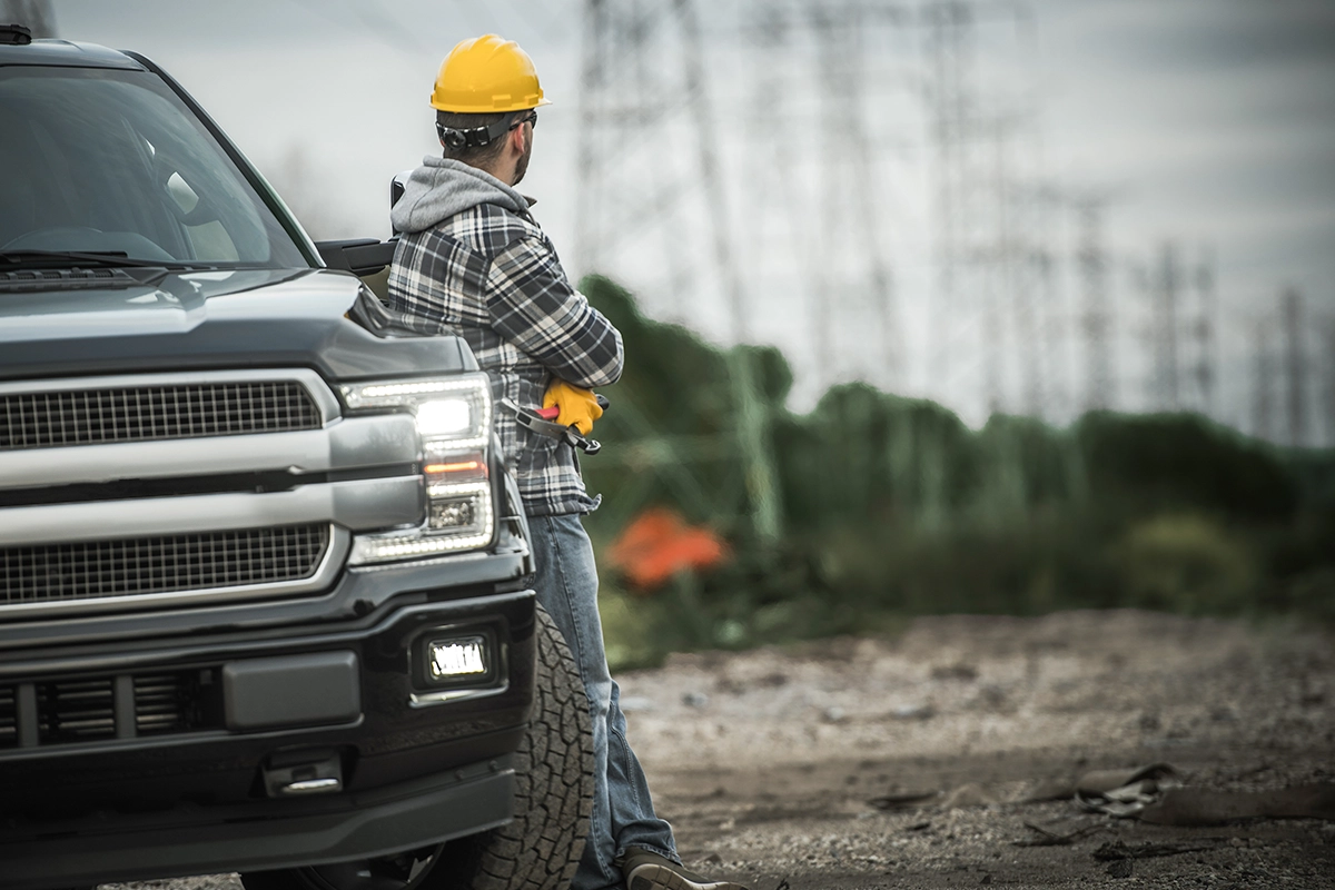 A construction worker in a yellow hard hat leaning against a black pickup truck with bright headlights
