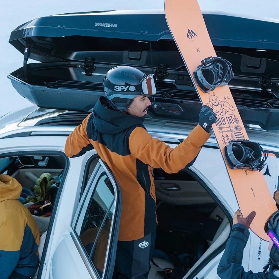 A man loading skis into a Yakima CBX XXL enclosed roof-mounted cargo box attached to a white SUV in the snow